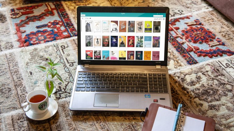 اپلیکیشن طاقچه در لپتاپ روی فرش ایرانی و کنار فنجان چای